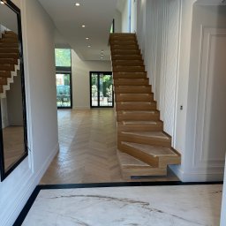 Usługi Budowlane Rafał Szczepanik - Jasne wnętrze domu z drewnianymi schodami w trakcie montażu, parkietem w jodełkę i marmurową podłogą. Widoczne listwy przypodłogowe i drzwi balkonowe.