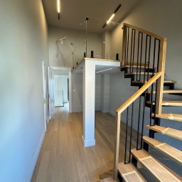 Usługi Budowlane Rafał Szczepanik - Nowoczesne wnętrze z drewnianymi schodami z czarną balustradą i oświetleniem liniowym. Widoczny fragment antresoli z szklaną barierką i jasna podłoga.