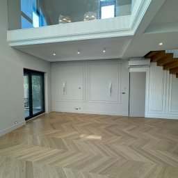Usługi Budowlane Rafał Szczepanik - Jasne, parkietowe podłogi ułożone we wzór jodełki francuskiej w przestronnym, nowocześnie wykończonym salonie z białymi ścianami i schodami.