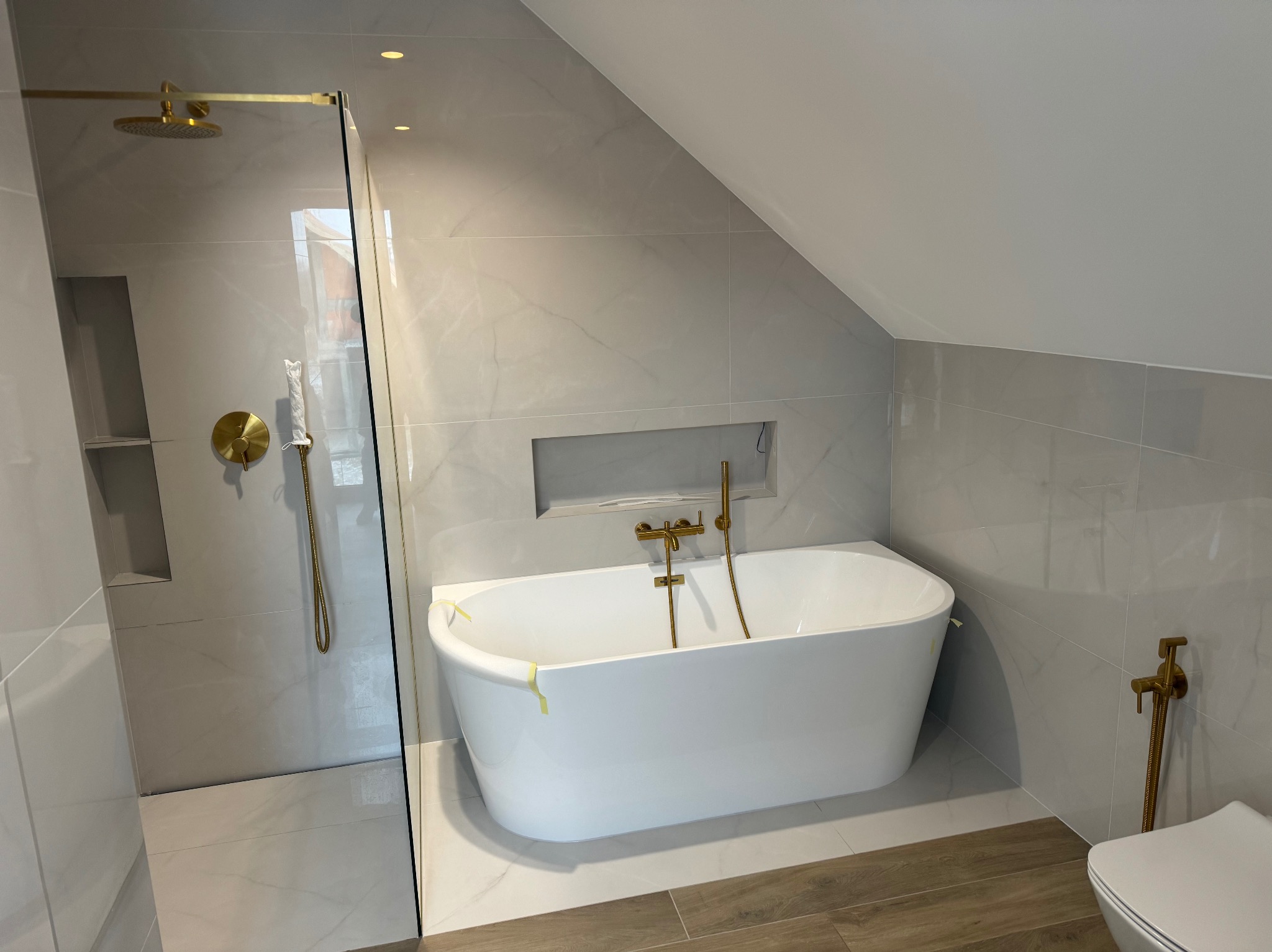 Nowoczesna łazienka na poddaszu z białą wanną wolnostojącą, złotą armaturą i kabiną prysznicową. Jasne płytki imitujące marmur na ścianach i podłodze.