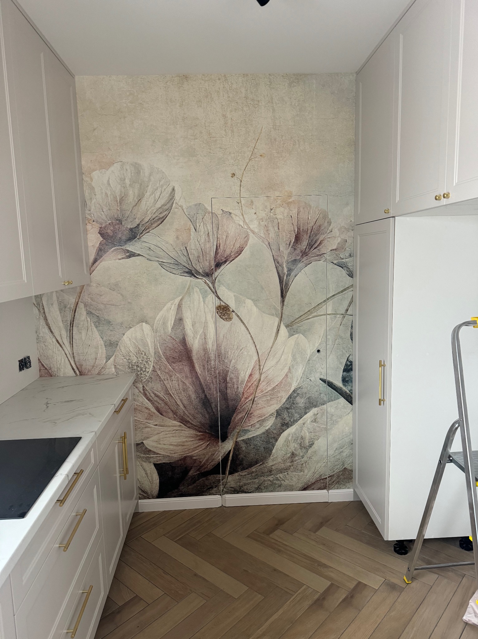 Biała zabudowa kuchenna z marmurowym blatem i złotymi uchwytami, na ścianie fototapeta z motywem kwiatowym, parkiet w jodełkę. Widoczna drabina.