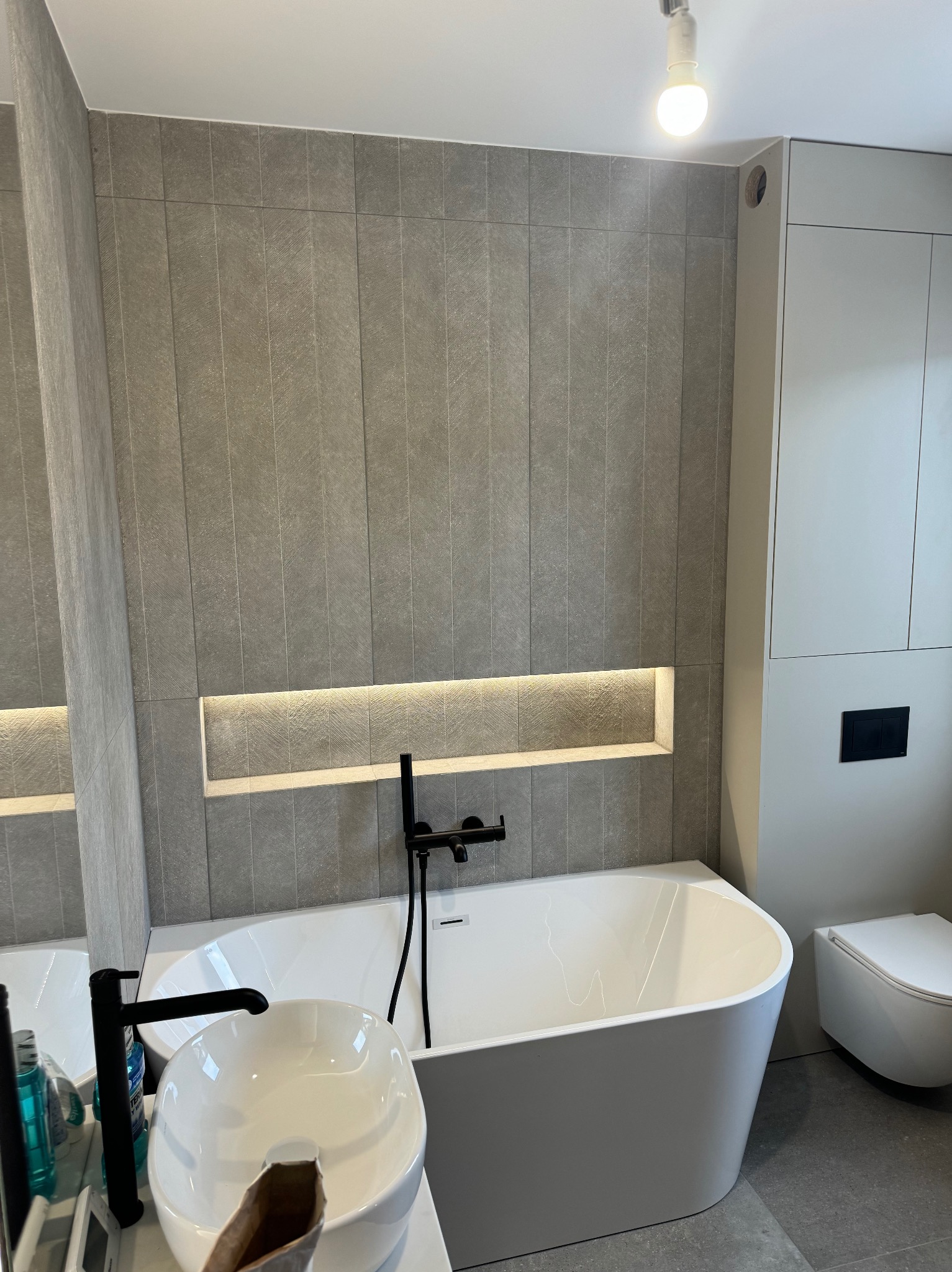 Nowoczesna łazienka z szarymi płytkami, podświetlaną wnęką, białą wanną i umywalką nablatową z czarną armaturą. Minimalistyczny design, stonowana kolorystyka.