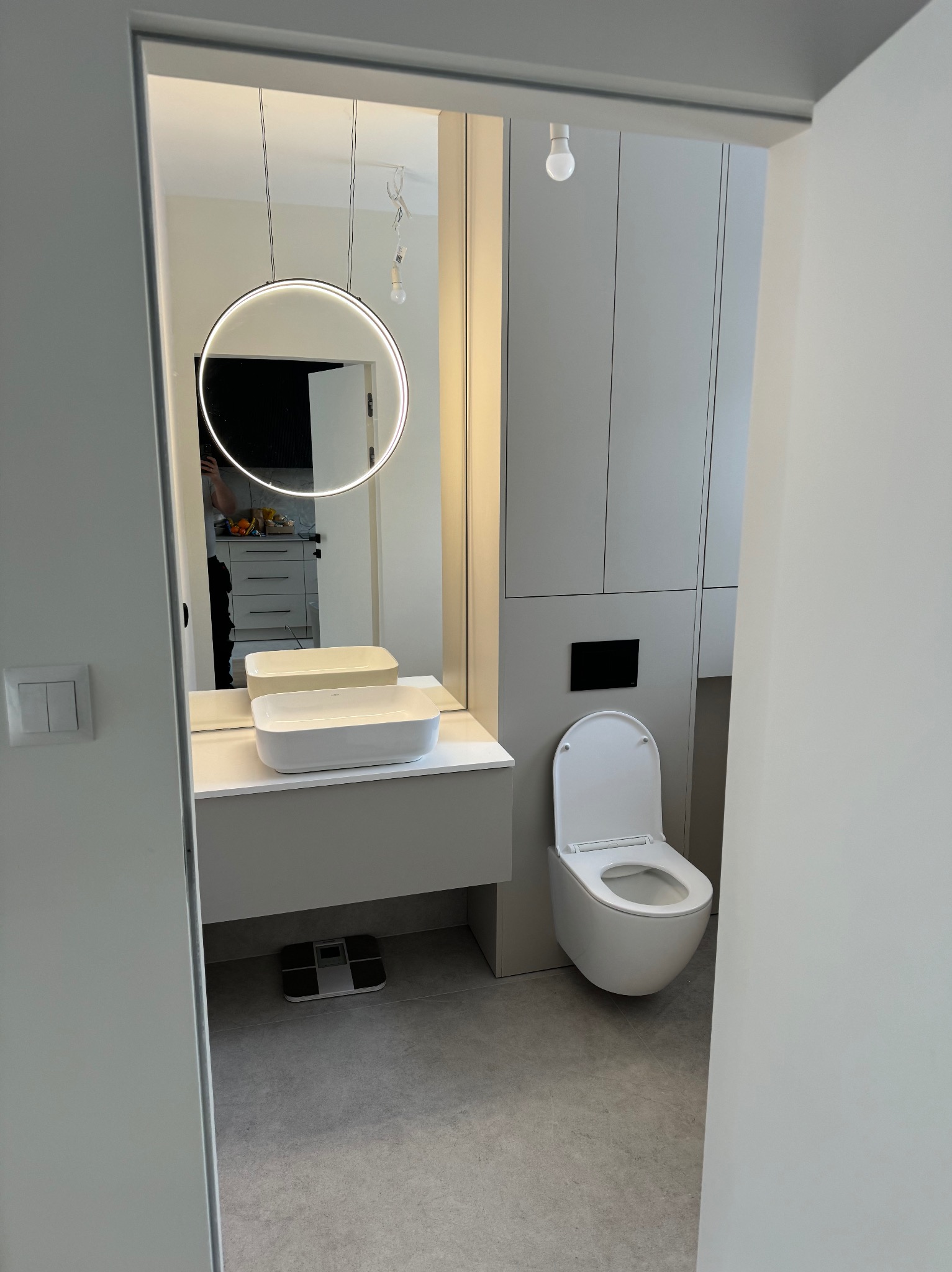 Nowoczesna łazienka w odcieniach szarości z podwieszaną toaletą i umywalką nablatową. W tle lustro z okrągłym oświetleniem LED i fragment kuchni.