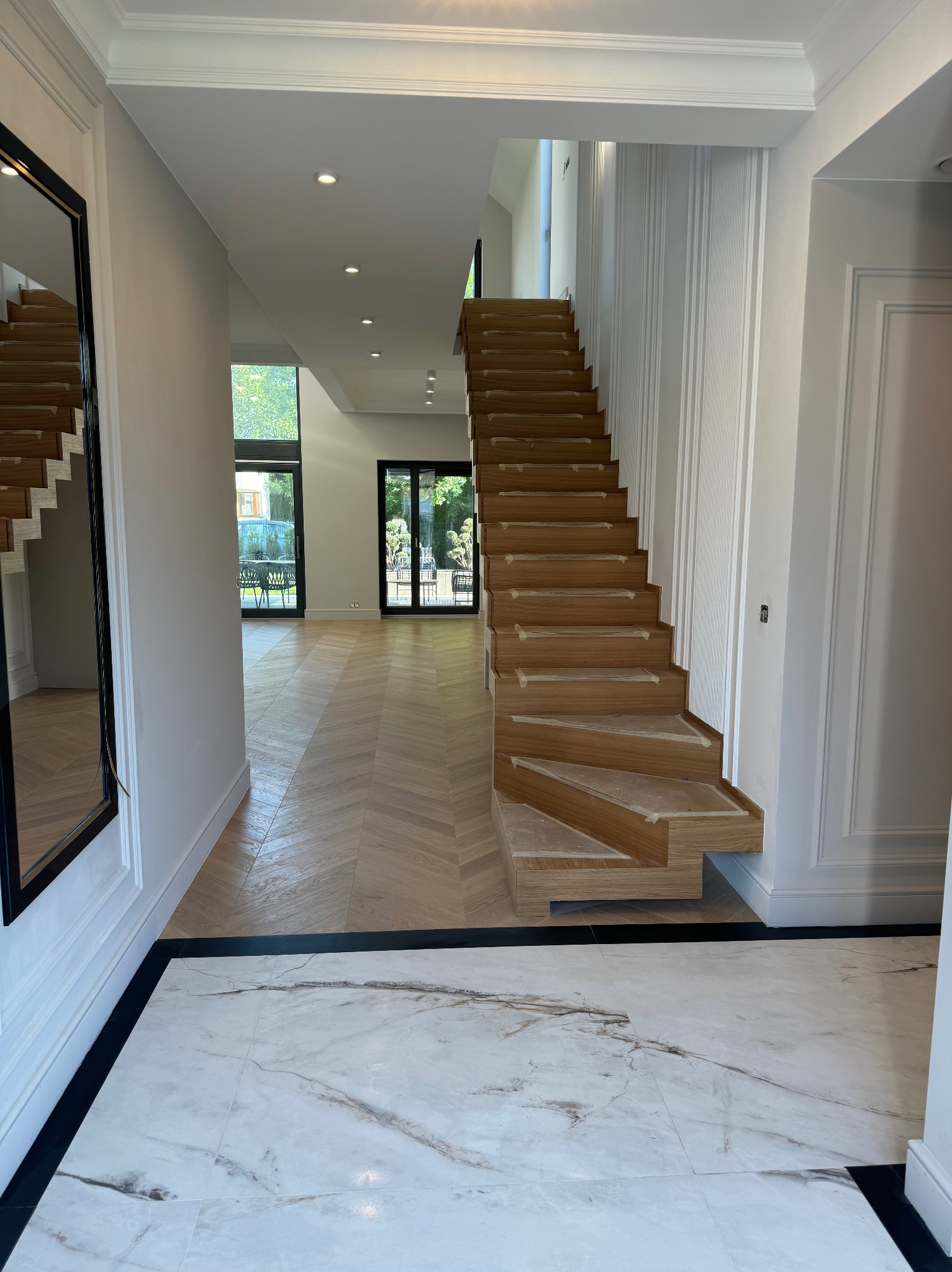 Jasne wnętrze domu z drewnianymi schodami w trakcie montażu, parkietem w jodełkę i marmurową podłogą. Widoczne listwy przypodłogowe i drzwi balkonowe.