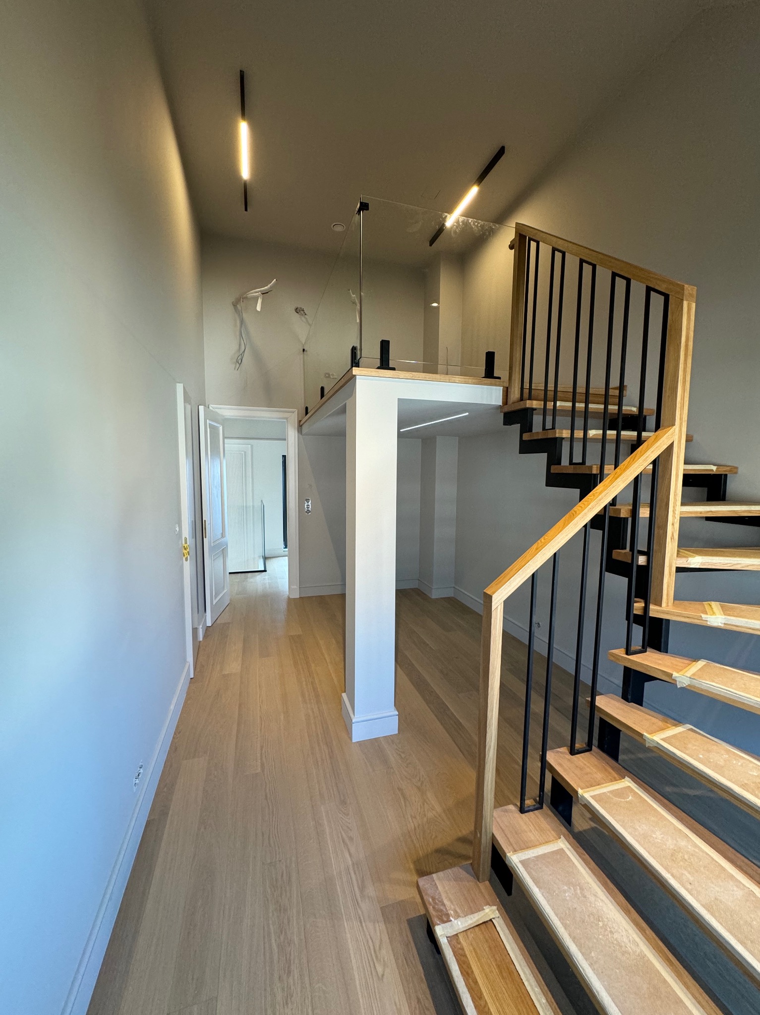 Nowoczesne wnętrze z drewnianymi schodami z czarną balustradą i oświetleniem liniowym. Widoczny fragment antresoli z szklaną barierką i jasna podłoga.