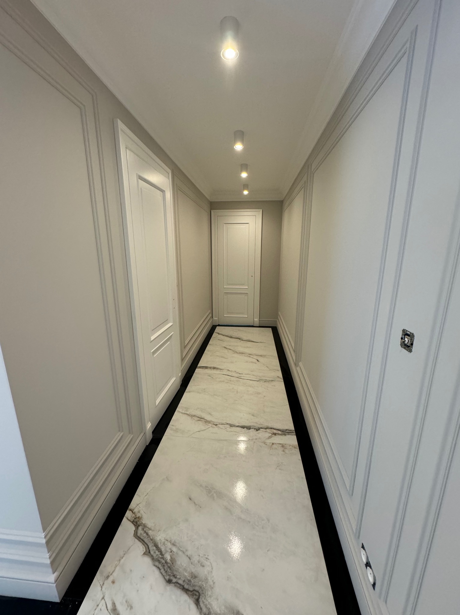 Korytarz po remoncie: białe ściany z dekoracyjnymi listwami, marmurowe płytki podłogowe, oświetlenie punktowe. Widok w perspektywie, z widocznymi drzwiami na końcu.