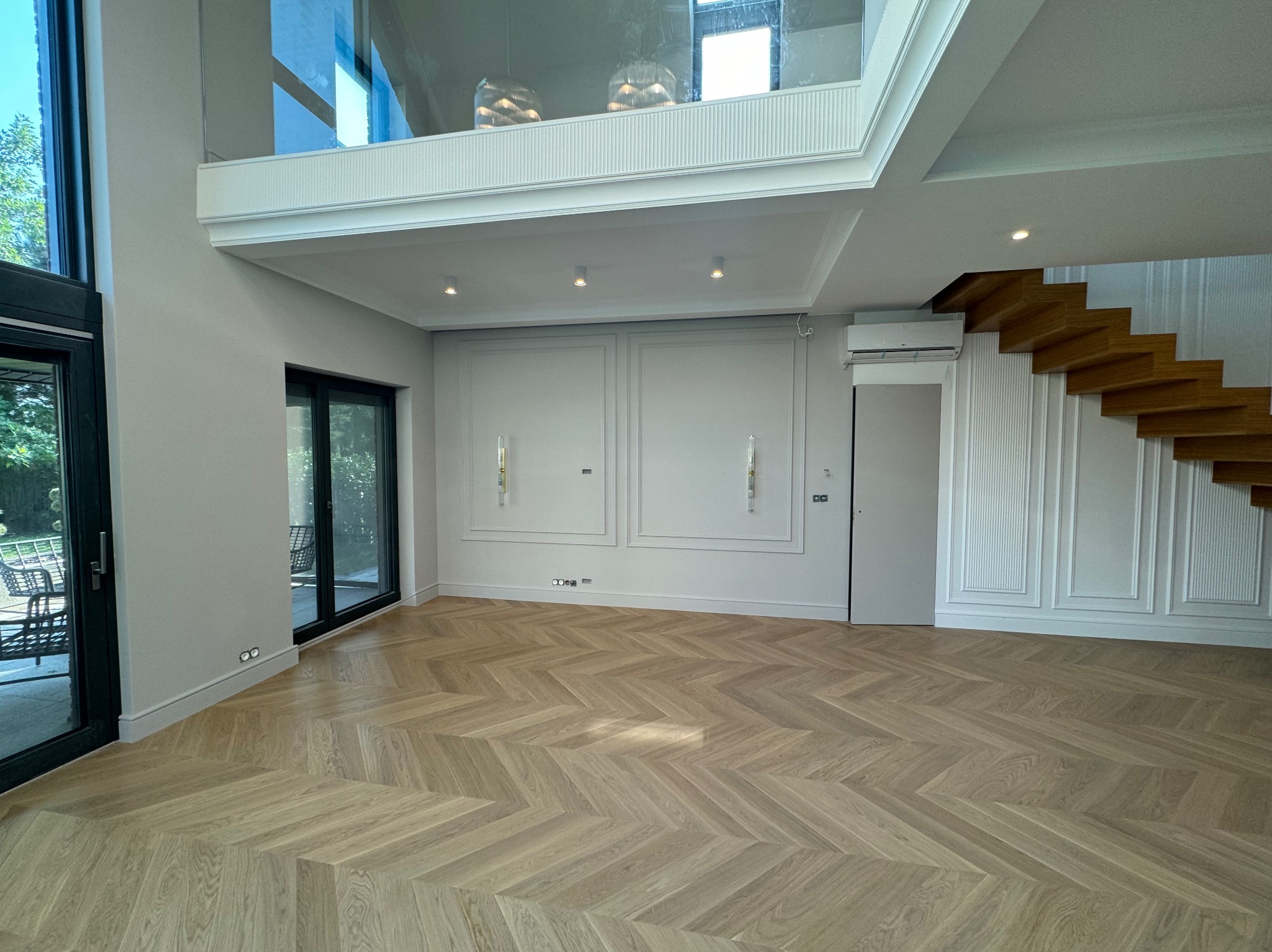 Jasne, parkietowe podłogi ułożone we wzór jodełki francuskiej w przestronnym, nowocześnie wykończonym salonie z białymi ścianami i schodami.