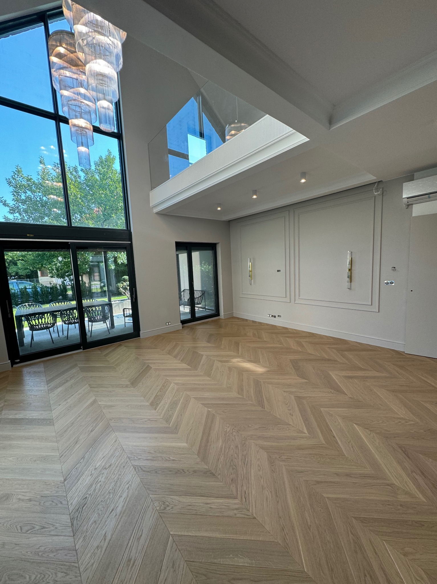 Jasny parkiet ułożony we wzór jodełki francuskiej w przestronnym salonie z wysokim oknem i antresolą. Minimalistyczne ściany z dekoracyjnymi panelami i nowoczesnym oświetleniem.