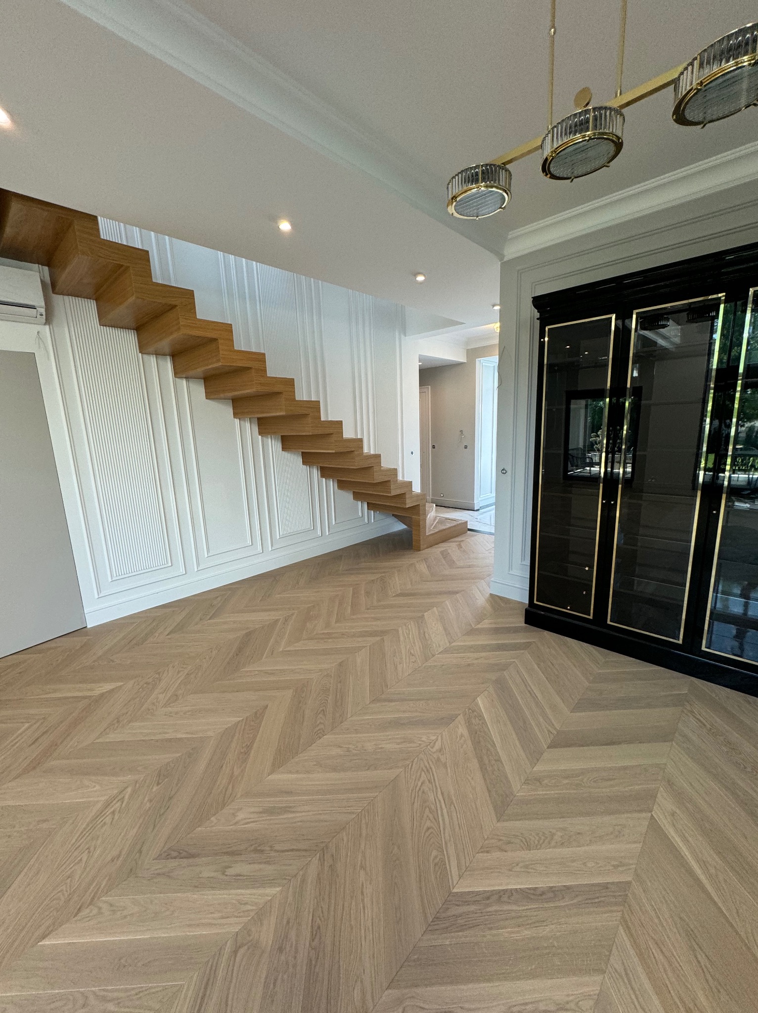 Nowoczesne wnętrze z drewnianymi schodami, parkietem w jodełkę i elegancką, czarną szafą z przeszkleniami. Minimalistyczny design z dbałością o detale.