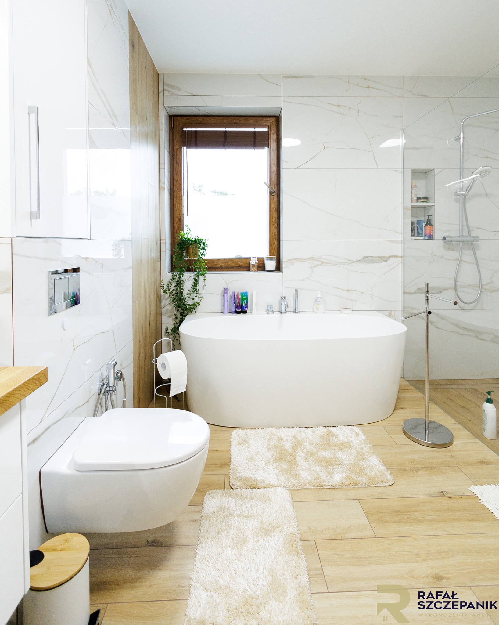 Jasna łazienka z wanną wolnostojącą, toaletą podwieszaną i drewnopodobnymi płytkami. Okno z drewnianą ramą i roletami. Minimalistyczny design z akcentami roślinnymi.