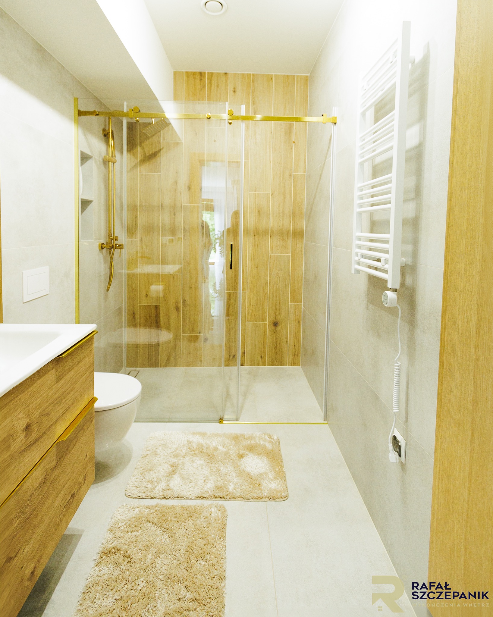 Jasna łazienka z kabiną prysznicową ze złotymi detalami, drewnianą szafką pod umywalką i grzejnikiem drabinkowym. Dwa beżowe dywaniki na podłodze.