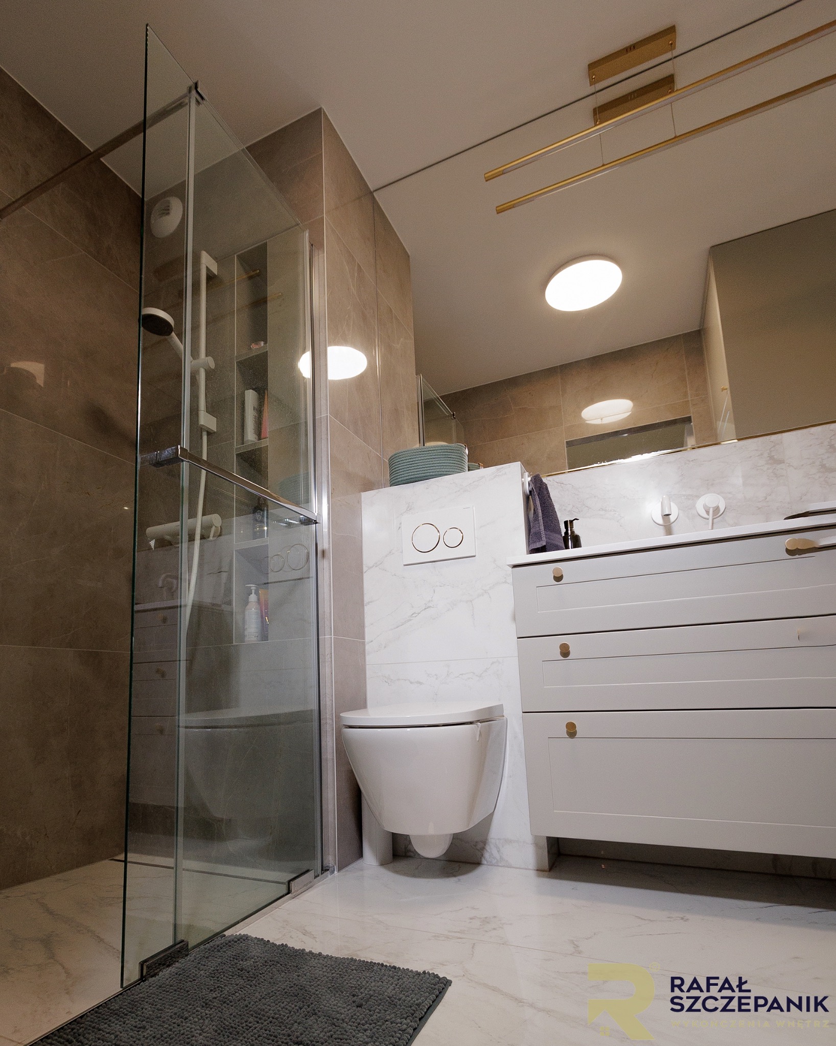 Nowoczesna łazienka z kabiną prysznicową typu walk-in, podwieszaną toaletą i szafką z umywalką. Beżowe płytki i marmurowa podłoga tworzą elegancki wystrój.