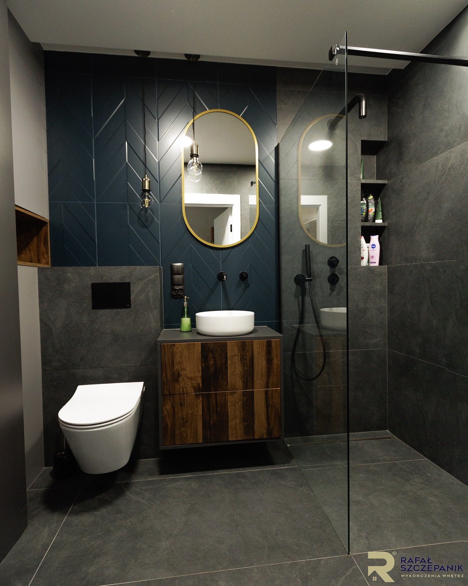 Nowoczesna łazienka z ciemnymi płytkami, kabiną prysznicową typu walk-in, owalnym lustrem w złotej ramie i drewnianą szafką pod umywalką. Minimalistyczny design.