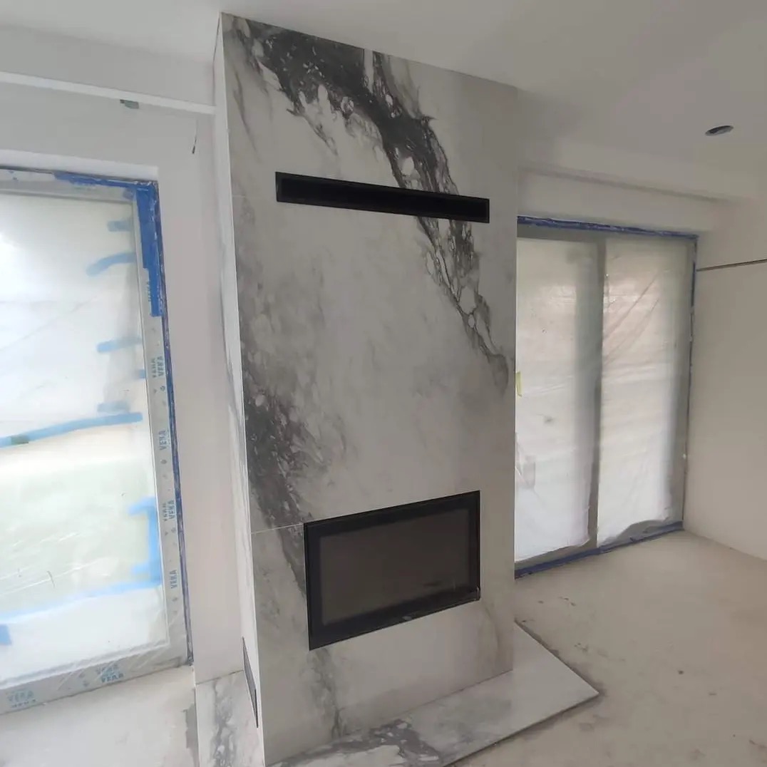 Nowoczesny kominek obłożony płytami imitującymi marmur z czarnym wkładem, w surowym wnętrzu z oknami zabezpieczonymi folią malarską.