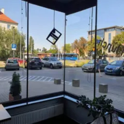 Widok z wnętrza lokalu przez duże, czyste okna na parking i ulicę w słoneczny dzień. Na szybach widoczne odbicia napisów. Minimalistyczna lampa wisi na tle nieba.