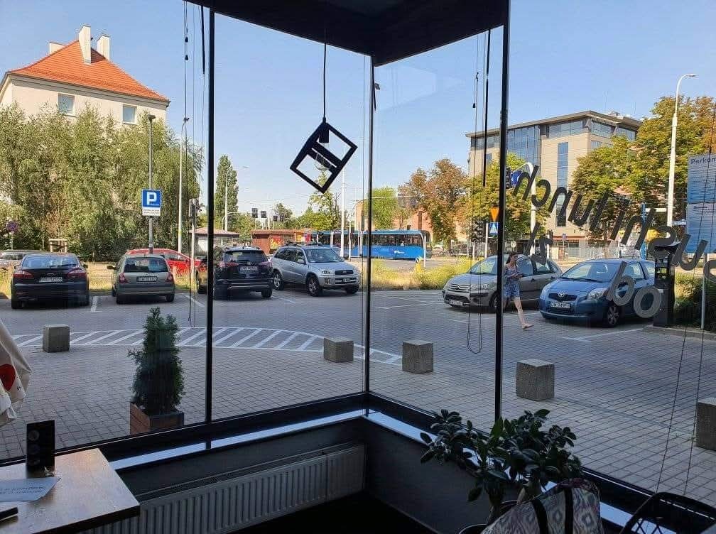 Widok z wnętrza lokalu przez duże, czyste okna na parking i ulicę w słoneczny dzień. Na szybach widoczne odbicia napisów. Minimalistyczna lampa wisi na tle nieba.