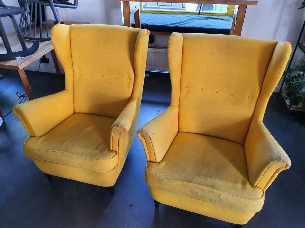 Dwa żółte fotele typu uszak, ustawione obok siebie na tle pokoju. Widoczne ślady użytkowania na siedziskach.