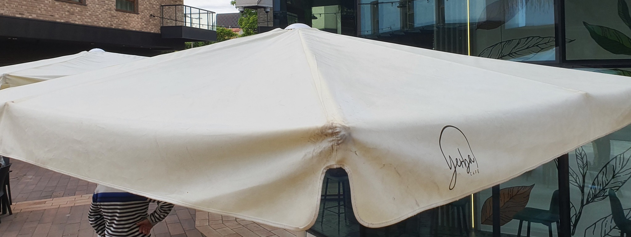 Biały, zabrudzony parasol kawiarniany z logo 'Yehba Cafe' na tle ceglanej ściany i przeszklonej elewacji. Widoczne ślady użytkowania.