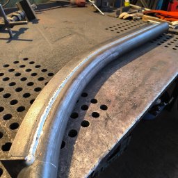 Well Welding Maciej Machulec - Wygięty, spawany element metalowy na perforowanym stole warsztatowym. Widoczne ślady spawania i narzędzia w tle. Surowy, przemysłowy klimat warsztatu.