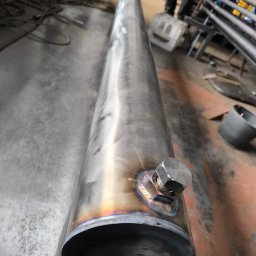 Well Welding Maciej Machulec - Metalowa rura z przyspawanym elementem, widoczne ślady po spawaniu. W tle elementy konstrukcyjne i podłoga hali. Detal z bliska ukazuje precyzję wykonania.