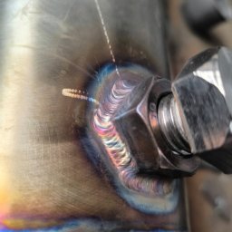 Well Welding Maciej Machulec - Zbliżenie na precyzyjne spawy łączące nakrętkę z metalową powierzchnią, ukazujące charakterystyczne, opalizujące zabarwienie termiczne.