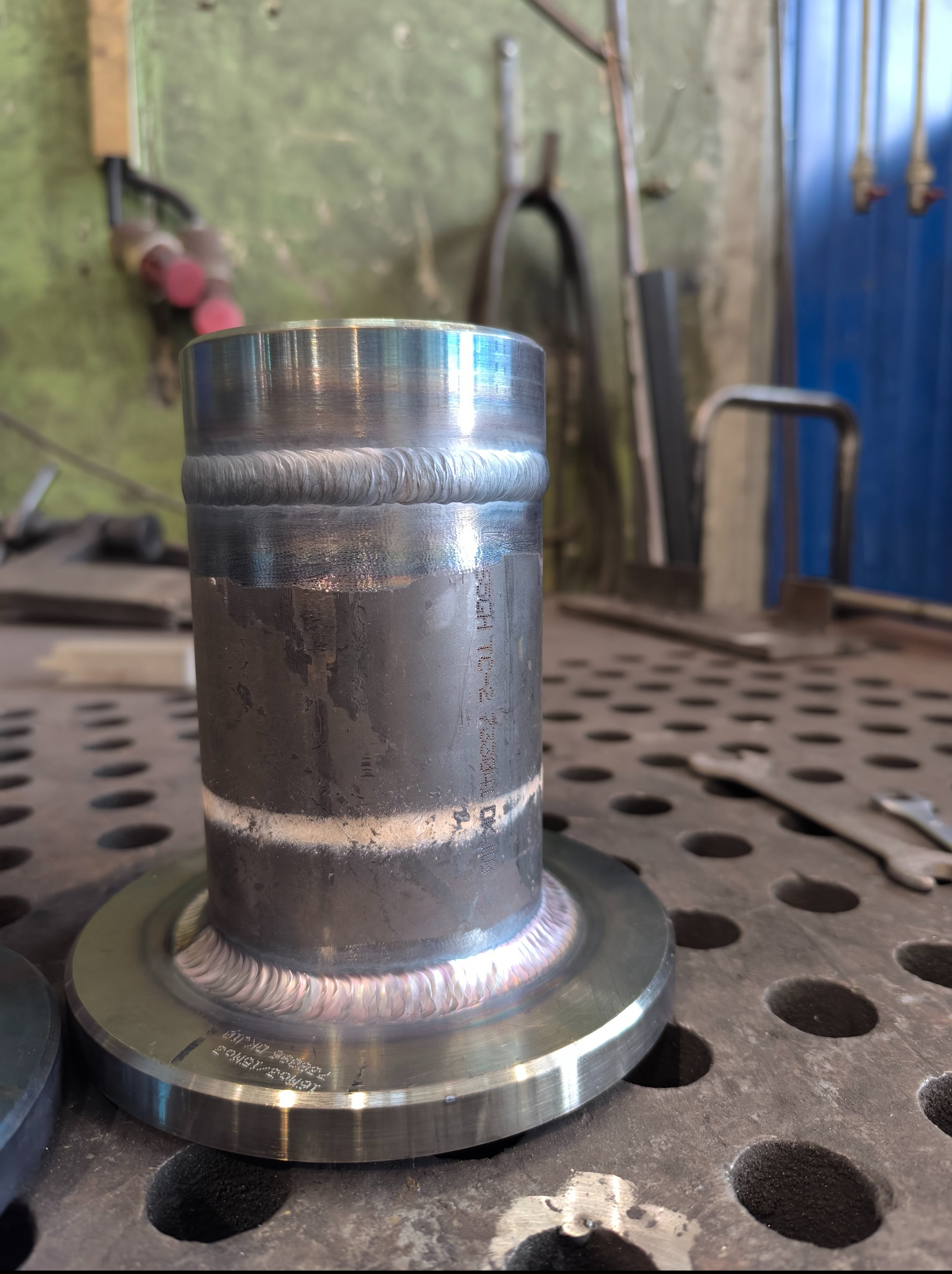 Metalowy element z widocznymi spawami, umieszczony na perforowanym stole warsztatowym. Widoczne narzędzia w tle i precyzyjne spoiny na obwodzie konstrukcji.