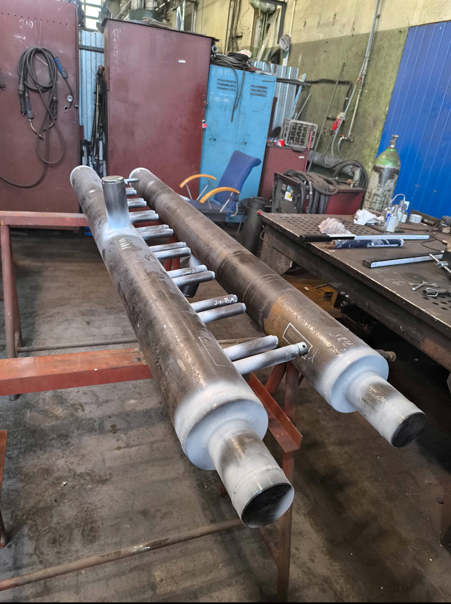 Dwa metalowe cylindry z przyspawanymi rurkami na metalowym stojaku w warsztacie. Widoczne ślady obróbki i spawania, tło stanowią narzędzia i wyposażenie warsztatowe.
