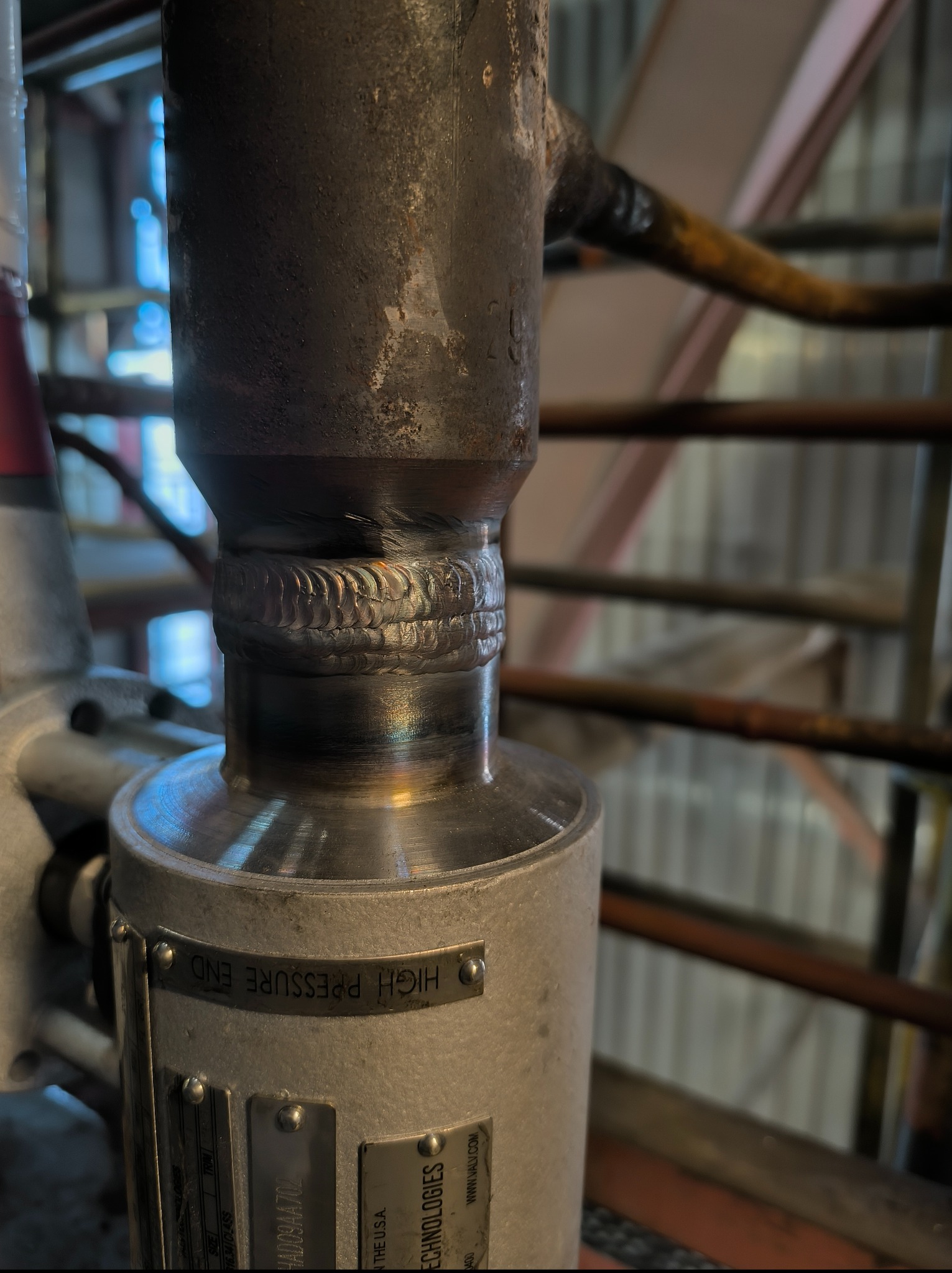 Metalowa konstrukcja z widocznym spawem, oznaczona tabliczką 'HIGH PRESSURE END', na tle schodów i elementów konstrukcyjnych.
