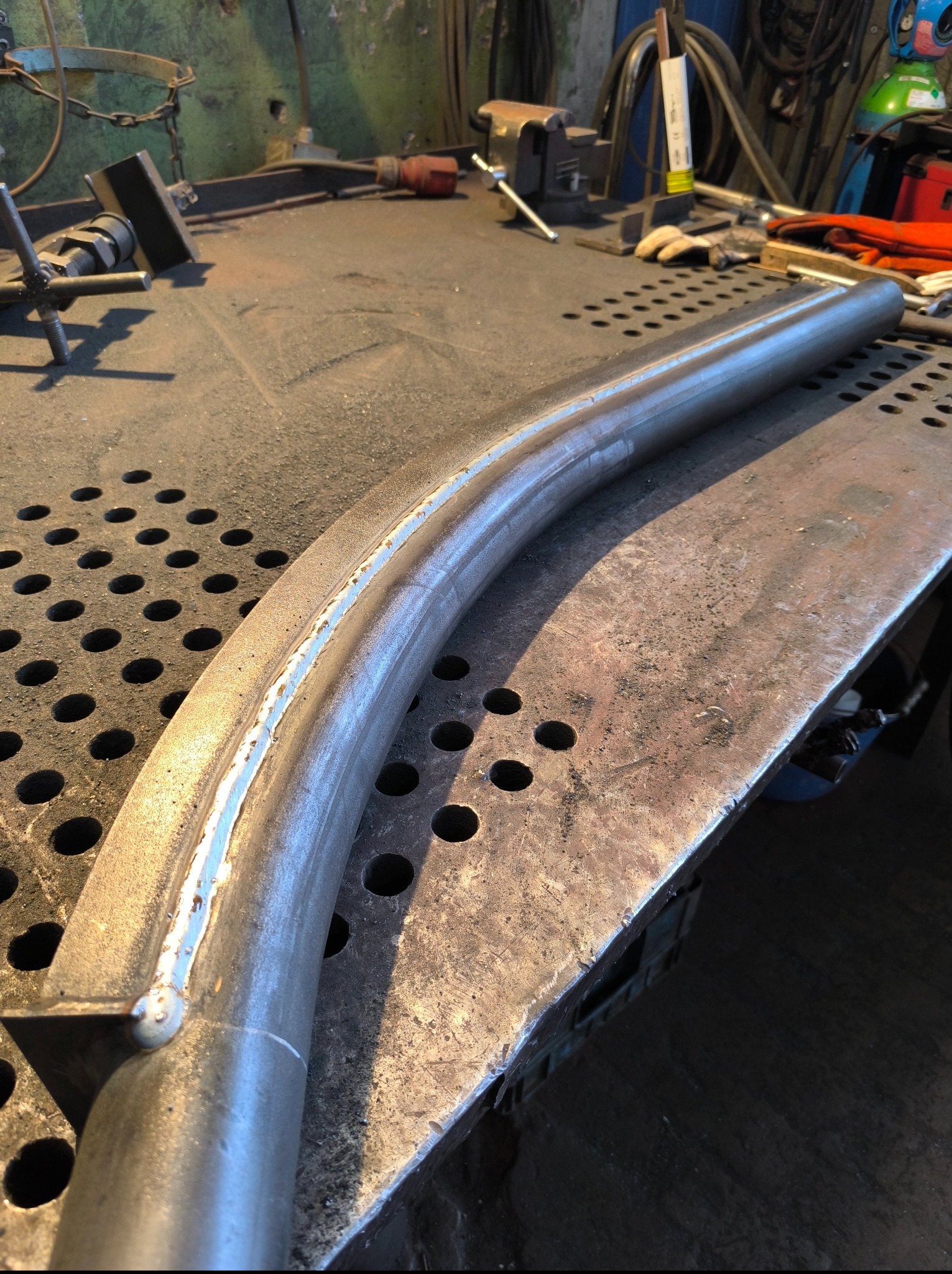 Wygięty, spawany element metalowy na perforowanym stole warsztatowym. Widoczne ślady spawania i narzędzia w tle. Surowy, przemysłowy klimat warsztatu.