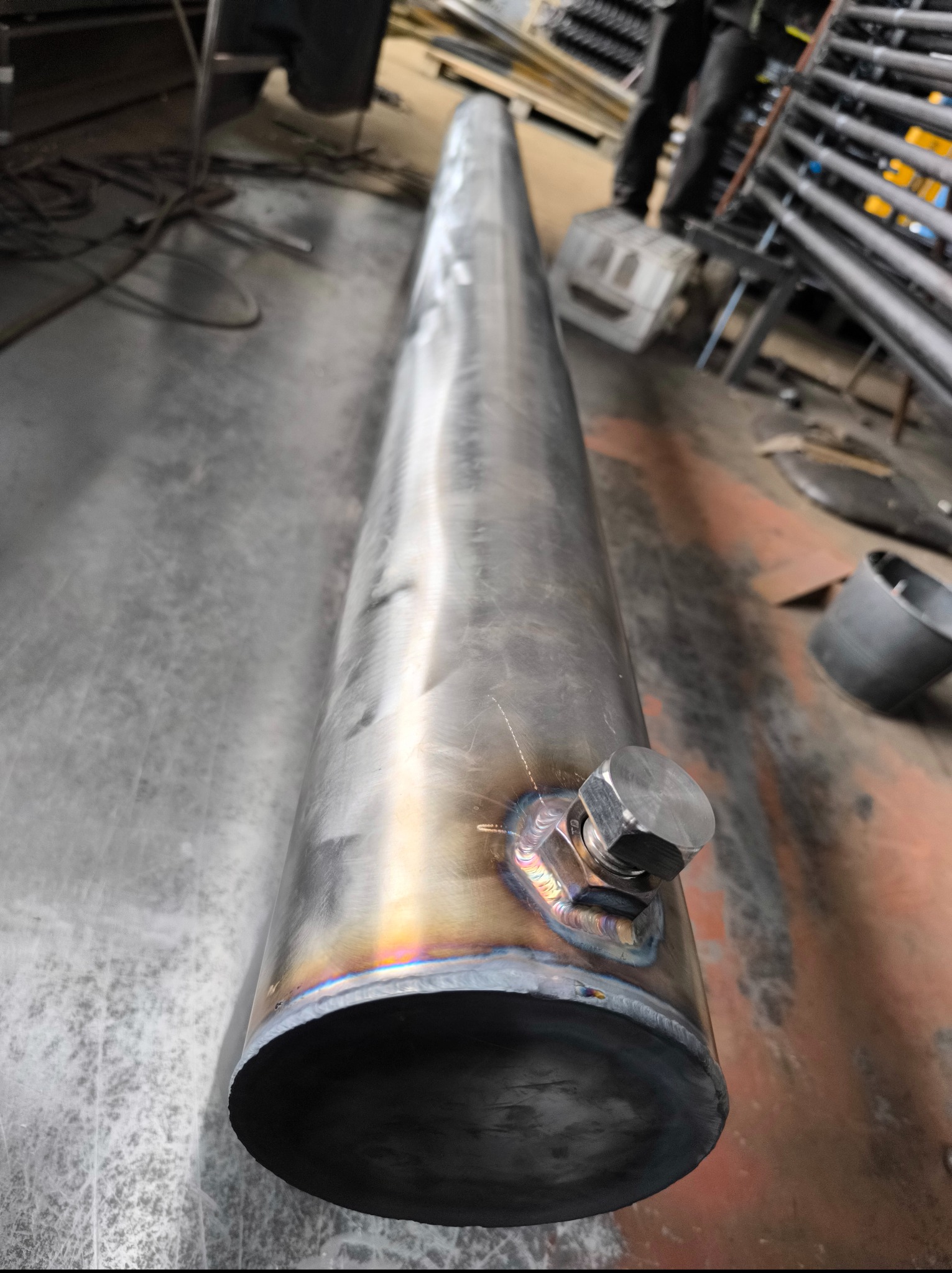 Metalowa rura z przyspawanym elementem, widoczne ślady po spawaniu. W tle elementy konstrukcyjne i podłoga hali. Detal z bliska ukazuje precyzję wykonania.