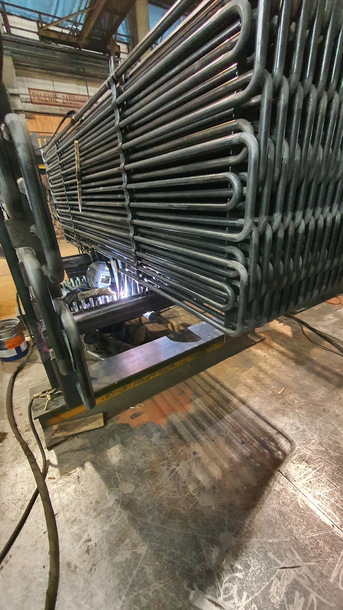 Proces łączenia metalowych rur przez spawanie. Widoczny spawacz w masce ochronnej, iskry i elementy konstrukcji. Ujęcie z bliska, oddające detale pracy.