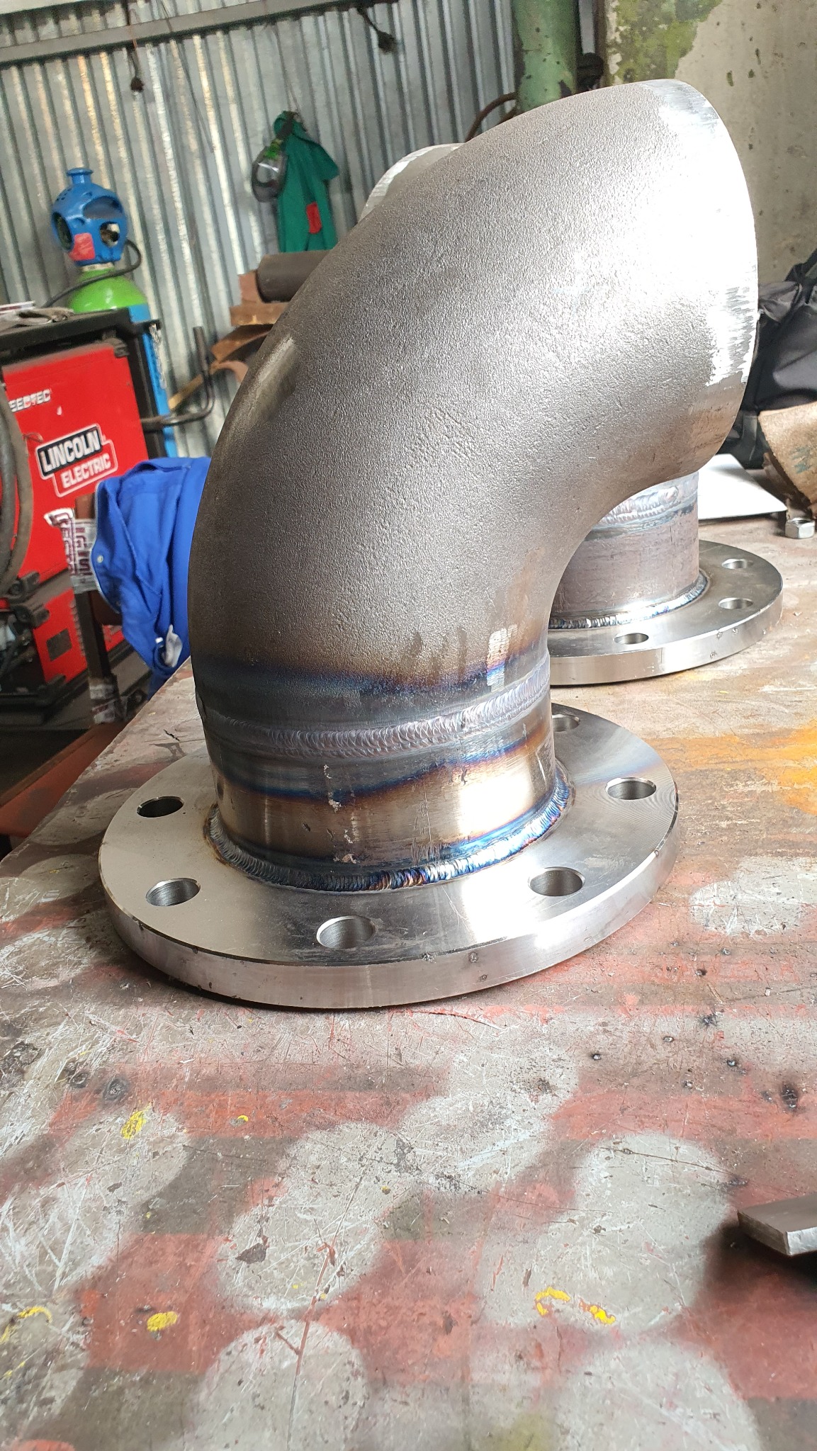 Dwa kolanka stalowe z przyspawanymi kołnierzami, widoczne ślady spawania, na stole warsztatowym z widocznym sprzętem spawalniczym w tle.