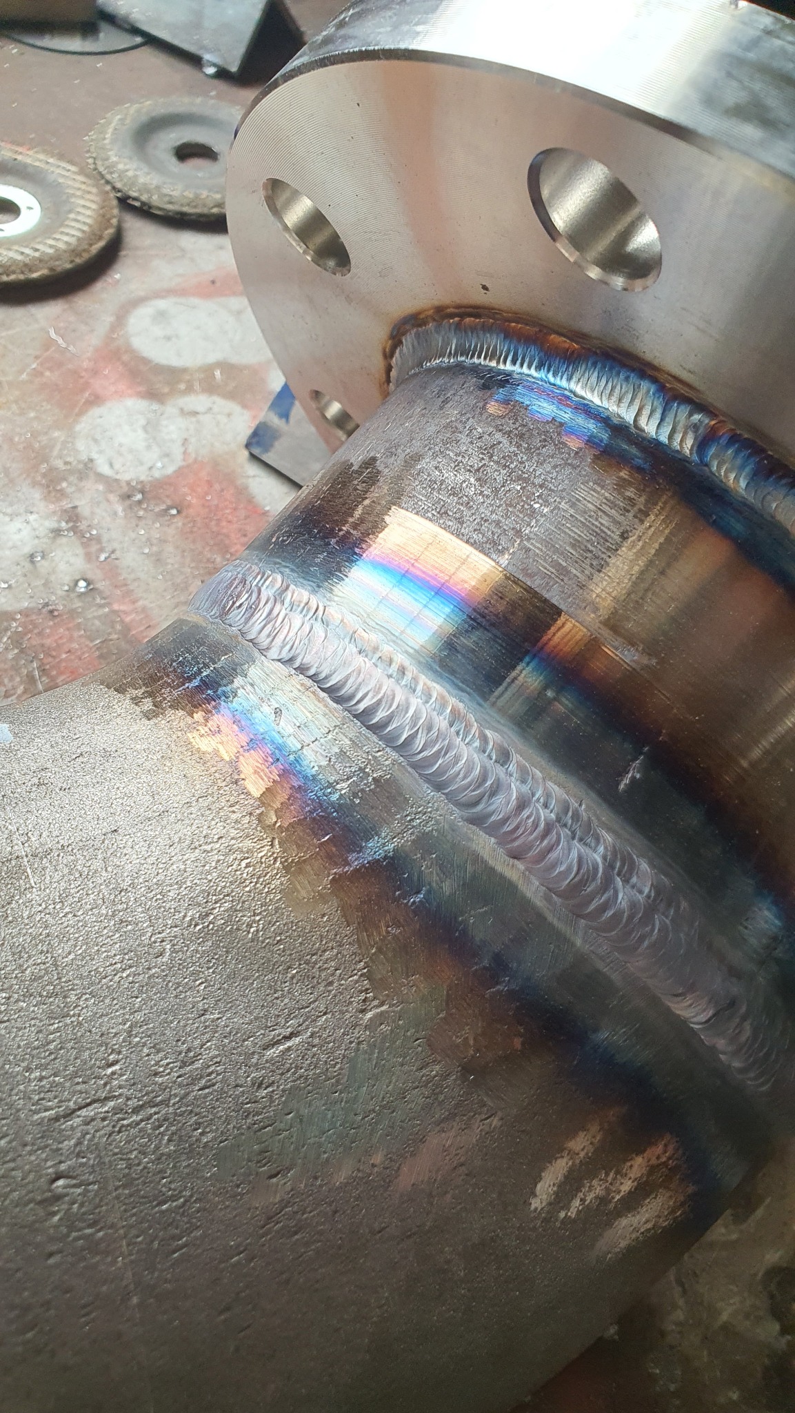 Precyzyjny spaw łączący rurę z kołnierzem, widoczne tęczowe przebarwienia po spawaniu. Szlifowane tło i tarcze szlifierskie w tle.