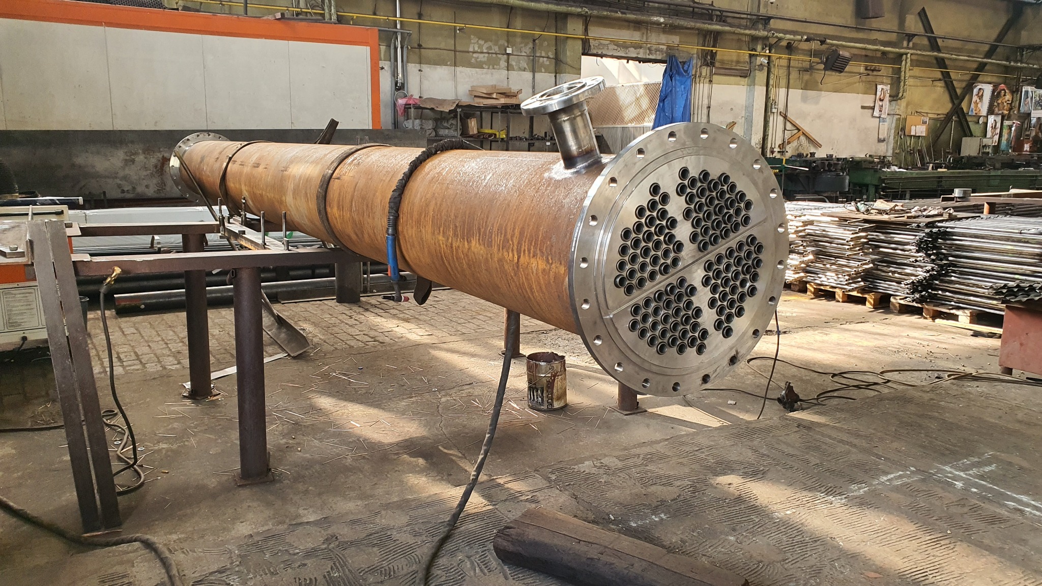 Skomplikowana konstrukcja metalowa z licznymi otworami, spawana w hali produkcyjnej. Widoczne ślady rdzy na cylindrycznym korpusie i elementy spawane, leżące na metalowym stojaku.
