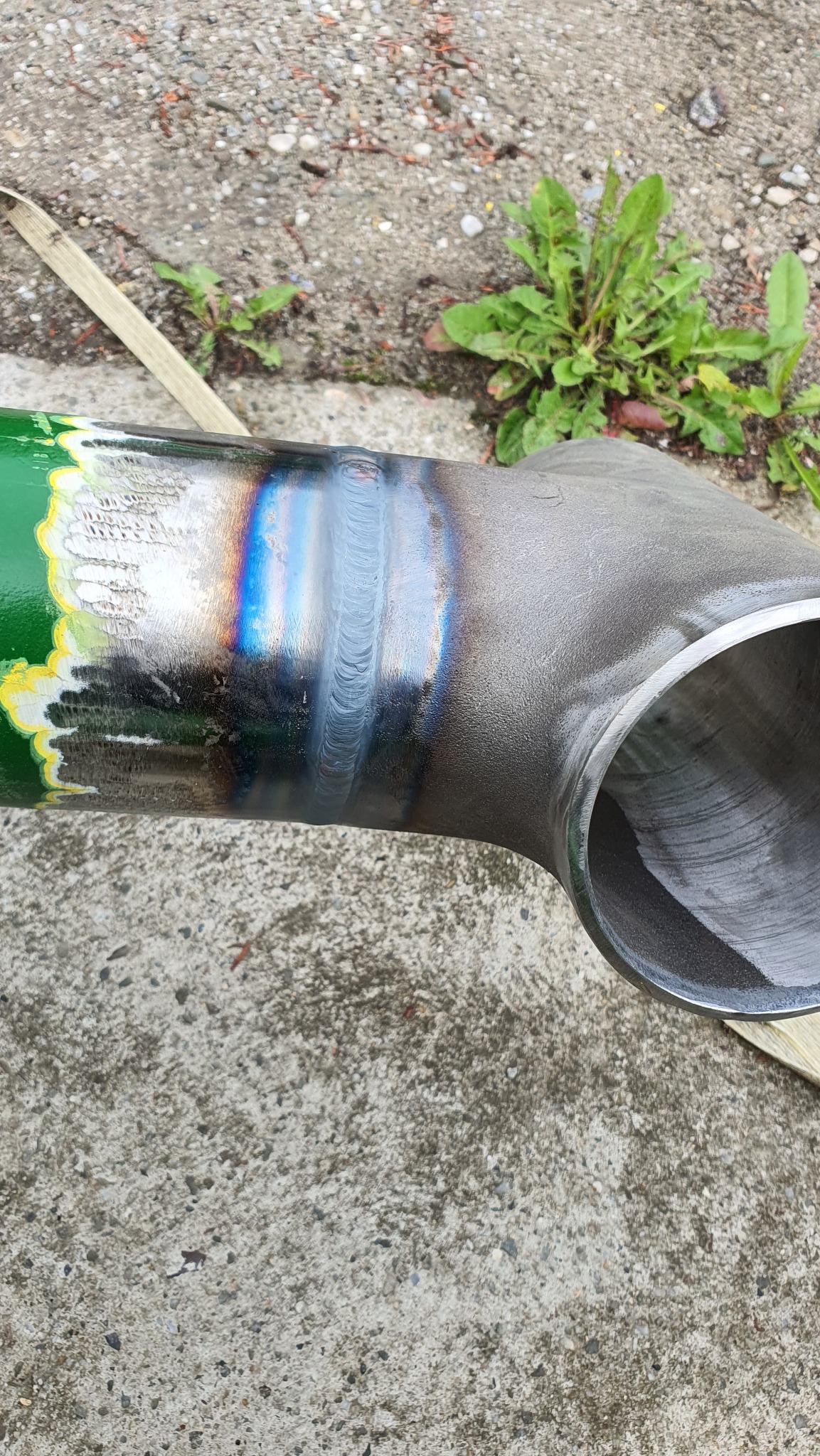 Spoina na metalowej rurze T-kształtnej, z widocznymi śladami obróbki i charakterystycznymi przebarwieniami termicznymi, leżącej na betonowym podłożu.