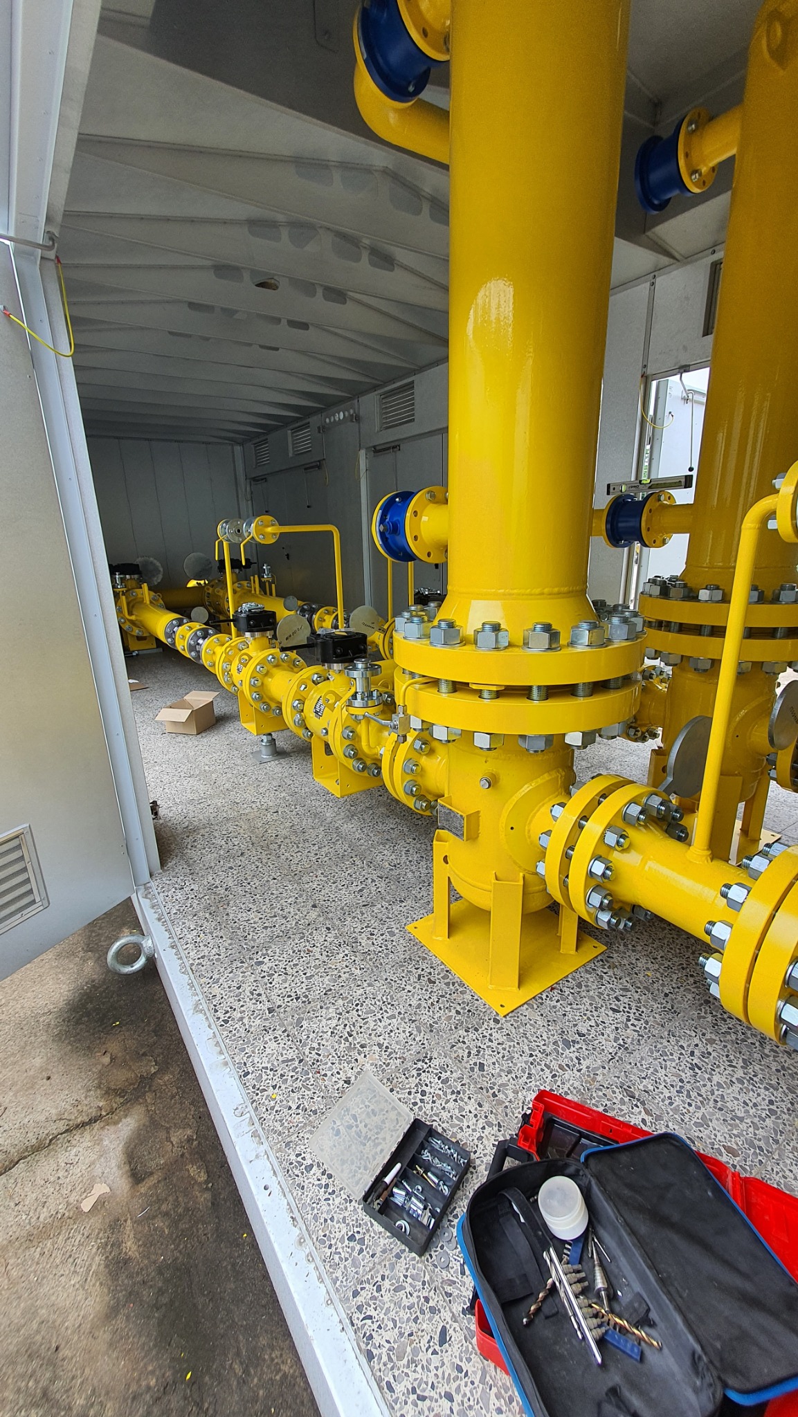 Żółta instalacja przemysłowa wewnątrz kontenera, widoczne liczne połączenia śrubowe i narzędzia ślusarskie. Elementy w kolorze niebieskim.