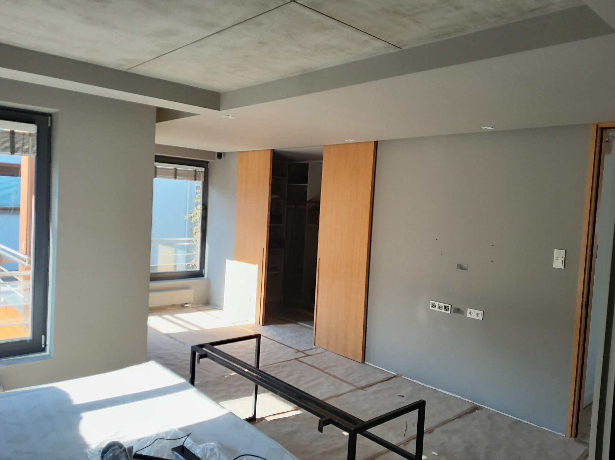 Nowoczesne wnętrze w trakcie wykończenia: szare ściany, drewniane drzwi przesuwne do garderoby, duże okna i surowy betonowy sufit. Widoczne przygotowania do dalszych prac.