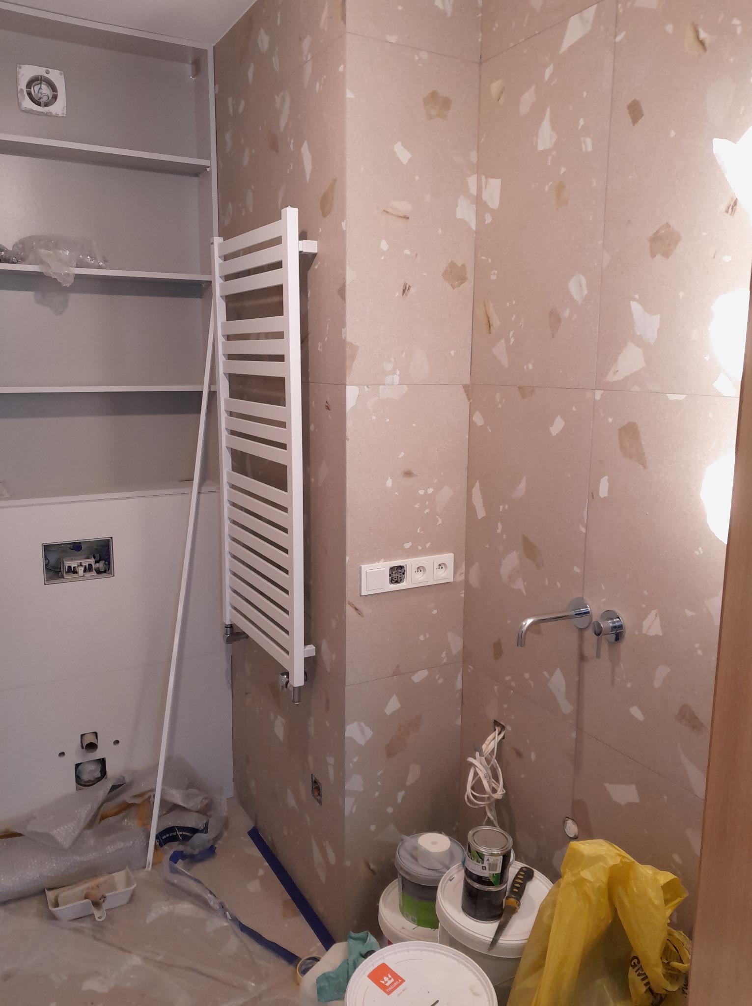 Remont łazienki w trakcie: biały grzejnik drabinkowy na ścianie z beżowych płytek, widoczne puszki z farbą i narzędzia, zabudowa WC ze stelażem podtynkowym.