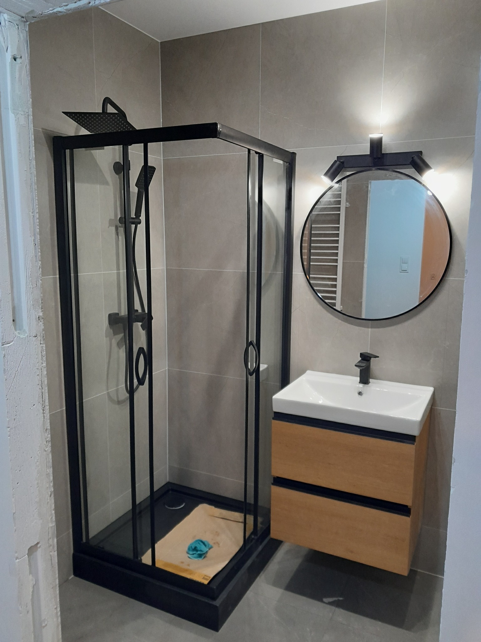 Nowoczesna łazienka w Białymstoku: kabina prysznicowa z czarną armaturą, umywalka z drewnianą szafką i okrągłe lustro z oświetleniem punktowym na szarych płytkach.