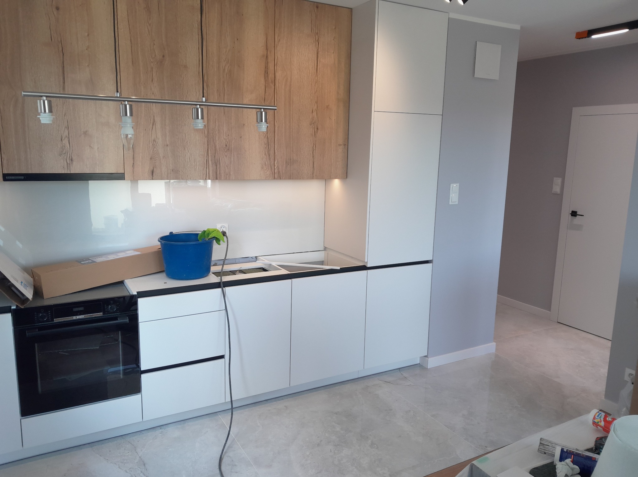 Nowoczesna kuchnia w trakcie montażu: jasne szafki z drewnianymi akcentami, sprzęt AGD, niebieskie wiadro i kartony na blacie, szare ściany, marmurowe płytki podłogowe.