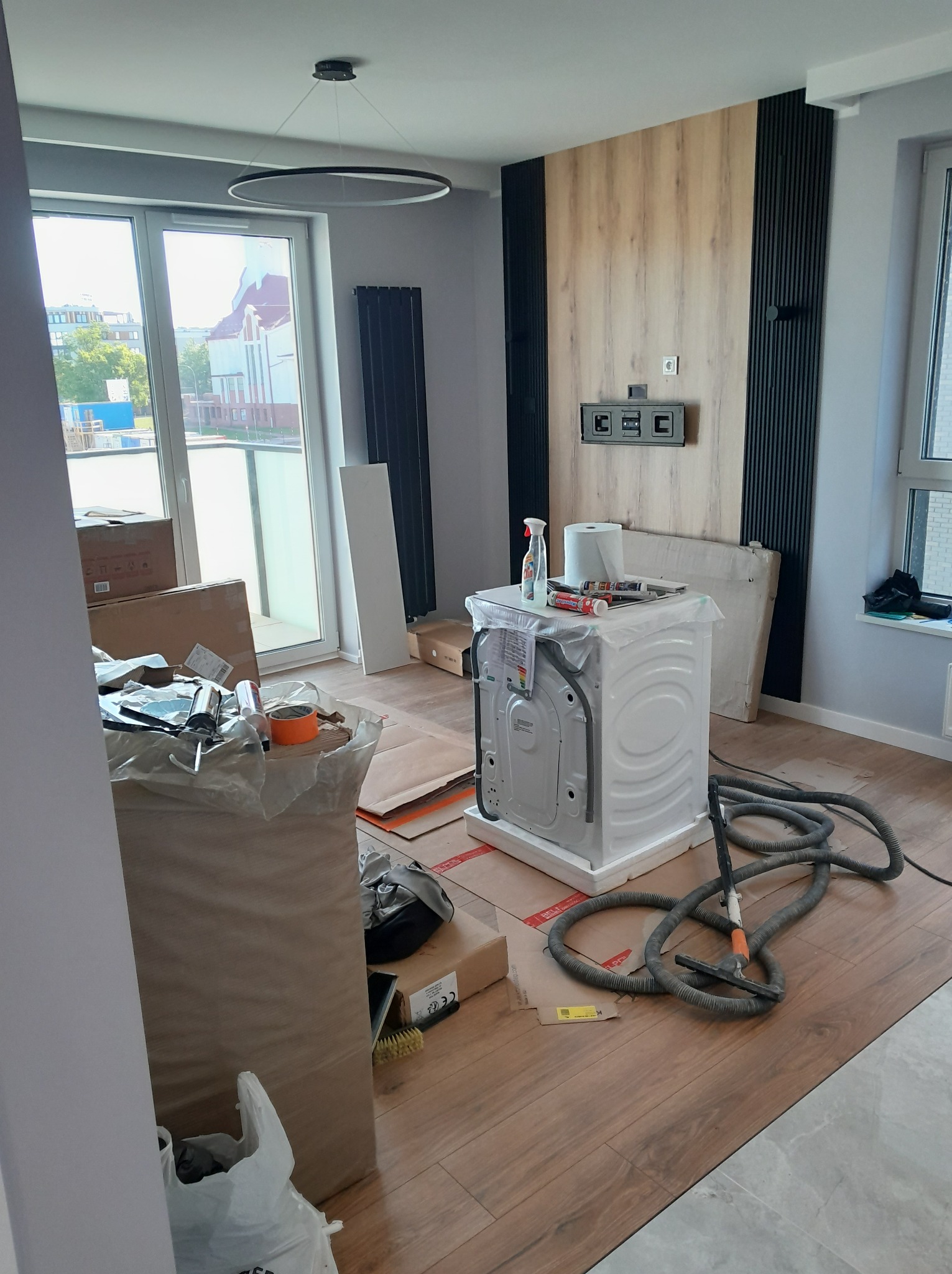 Wnętrze po remoncie: drewniana ściana z czarnymi panelami, nowa pralka, podłoga drewnopodobna. Widoczne narzędzia i materiały, sugerujące etap wykończeniowy.