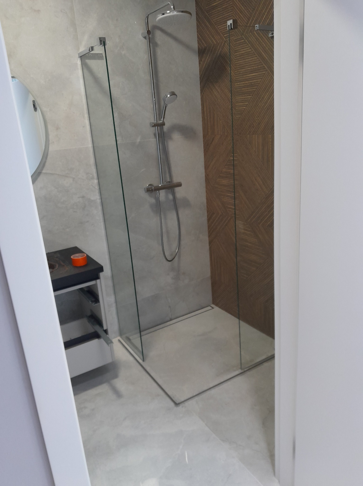 Nowoczesna łazienka po remoncie: kabina prysznicowa ze szklaną ścianką, szara marmurowa podłoga i ściany, umywalka z szafką. Widok zza futryny.