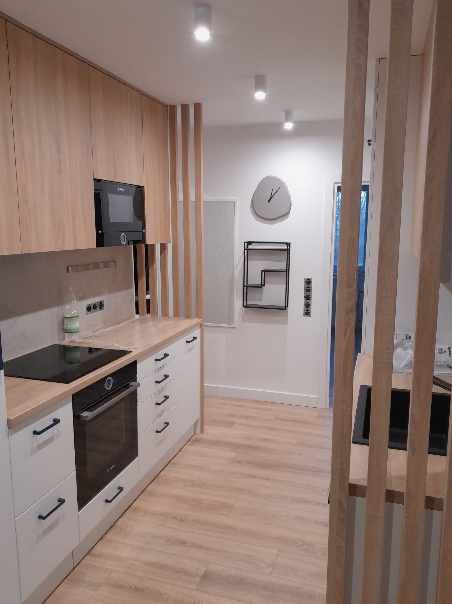 Jasna kuchnia z drewnianymi akcentami: zabudowa meblowa z piekarnikiem, płyta grzewcza, drewniana podłoga i dekoracyjna ścianka działowa z drewnianych listew.