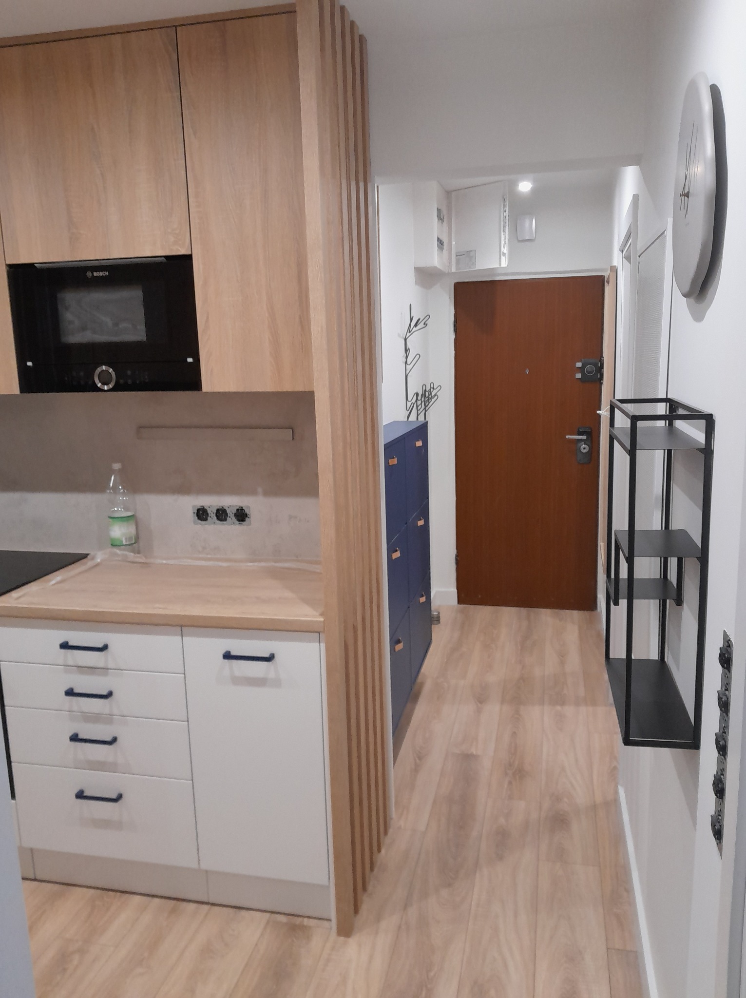 Jasna kuchnia z drewnianymi elementami i przedpokój z drewnianą podłogą. Widoczny czarny piekarnik Bosch, białe szafki z granatowymi uchwytami, niebieska szafka na buty.