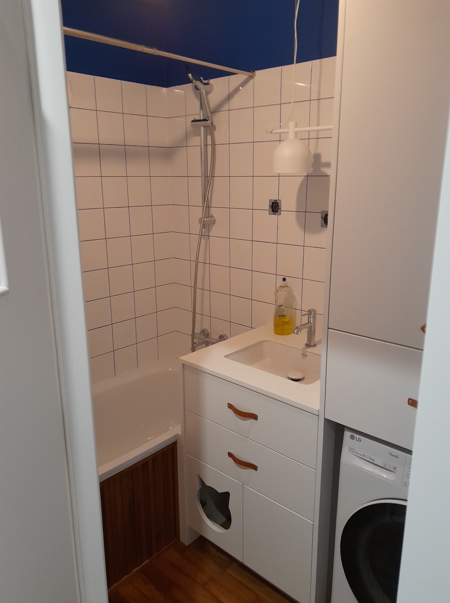 Biała łazienka z wanną, umywalką i pralką. Uwagę przyciąga niebieski sufit oraz szafka pod umywalką z wyciętym kocim kształtem. Widok zza framugi.