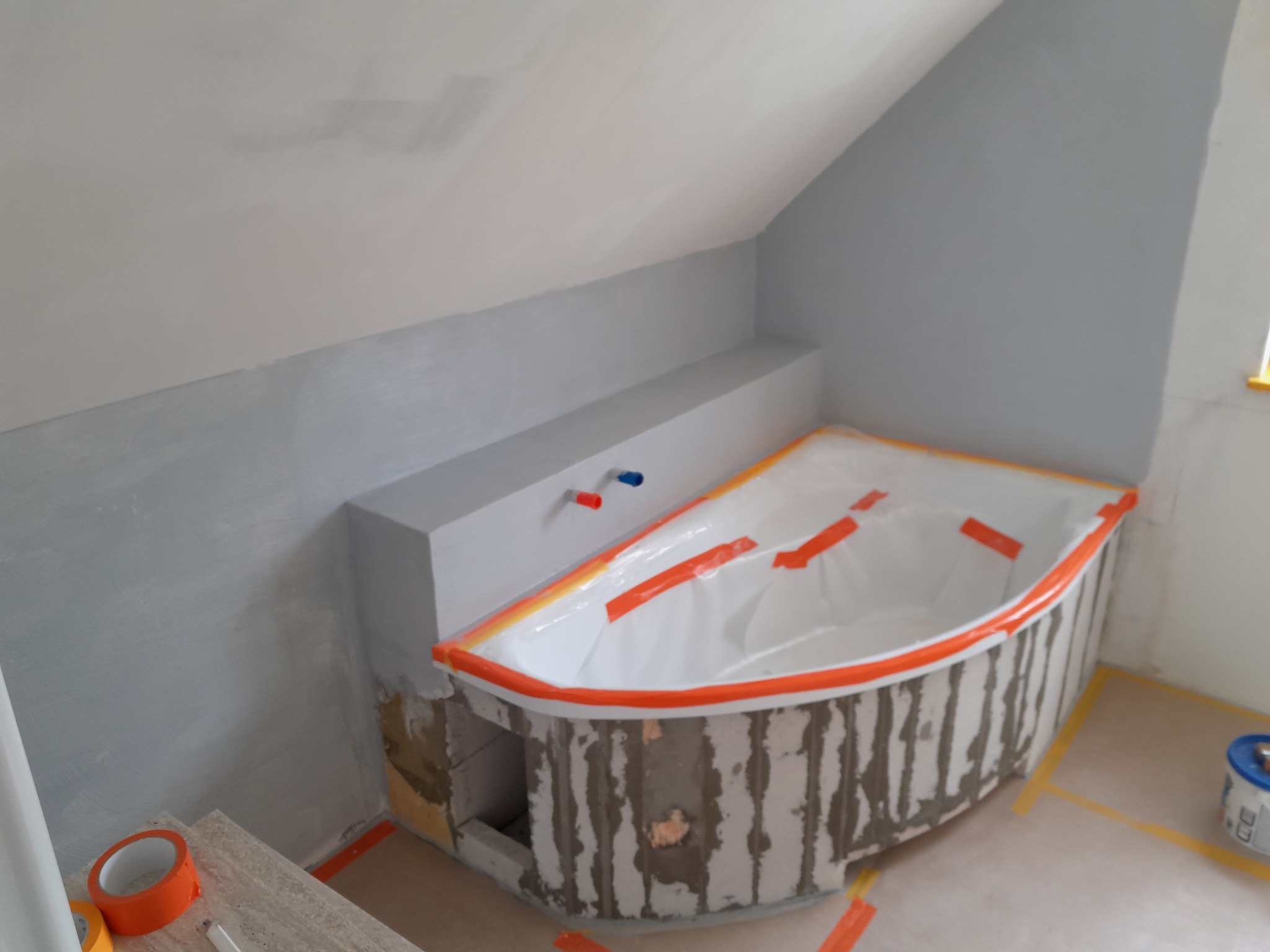 Remont łazienki na poddaszu: wanna obudowana bloczkami, zabezpieczona folią i taśmą. Ściany częściowo pomalowane na szaro, widoczne materiały budowlane.