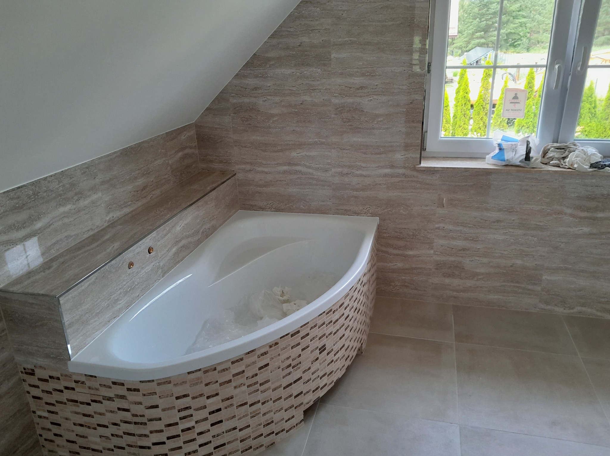 Wykończona łazienka na poddaszu: beżowe płytki imitujące marmur na ścianach, mozaika na obudowie wanny, biała wanna narożna, okno z widokiem na zieleń.