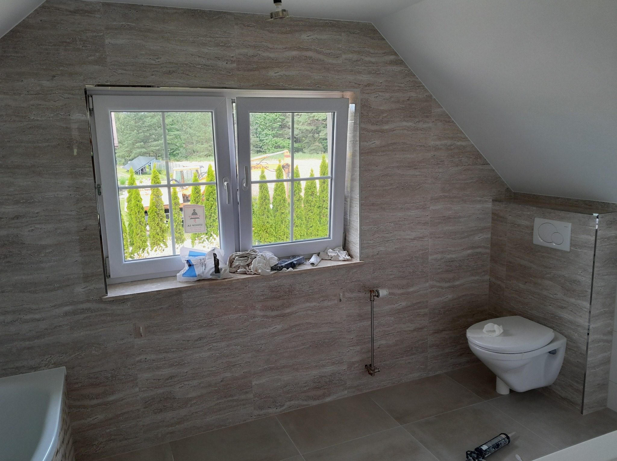 Ukończona łazienka na poddaszu z oknem, wykończona płytkami imitującymi trawertyn. Widoczny biały sedes podwieszany i fragment wanny. Na parapecie narzędzia i akcesoria.
