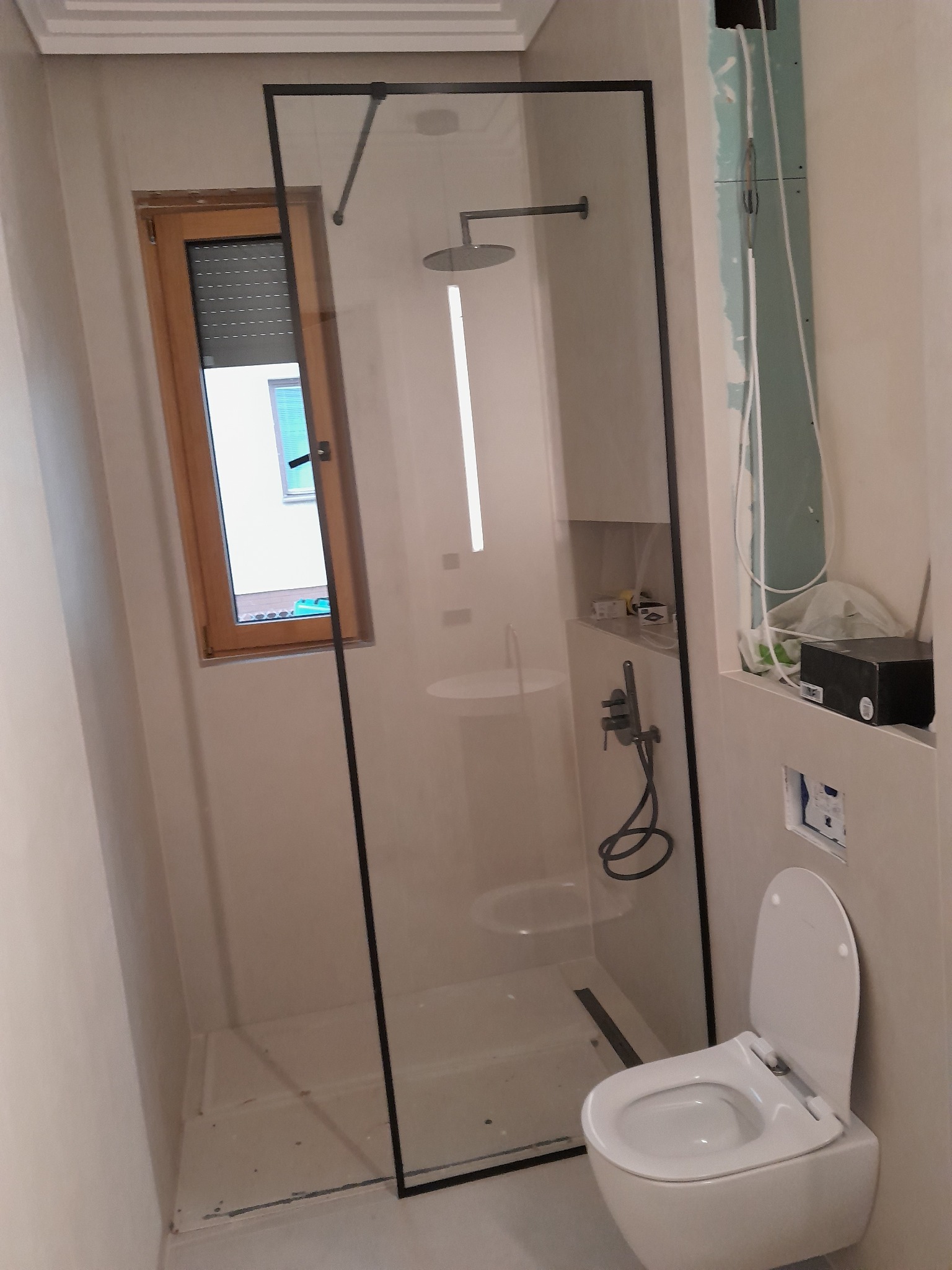 Nowoczesna kabina prysznicowa typu walk-in z czarnymi profilami, widoczna toaleta i okno z roletą w jasnej łazience w trakcie remontu. Ściany w jasnym odcieniu.