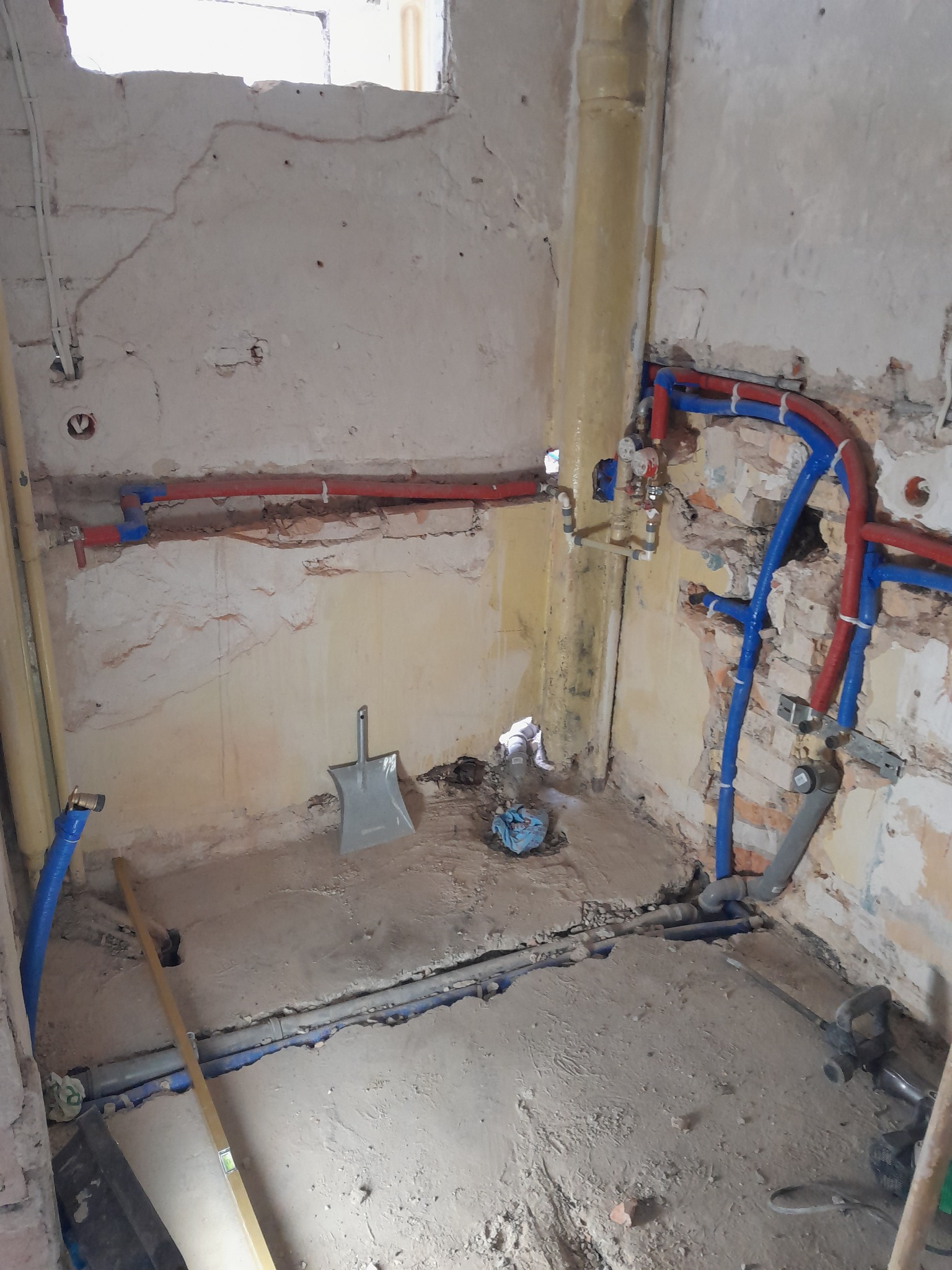 Łazienka w trakcie remontu: odkryte instalacje wodne (czerwone i niebieskie rury), kanalizacyjne i fragment ściany z widocznymi warstwami tynku. Widoczna łopata i poziomica.